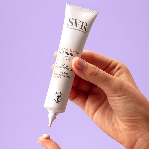 La mano de una mujer aplicando producto en uno de sus dedos del Creme SPF50+ de ClaIrial de SVR en un fondo gris