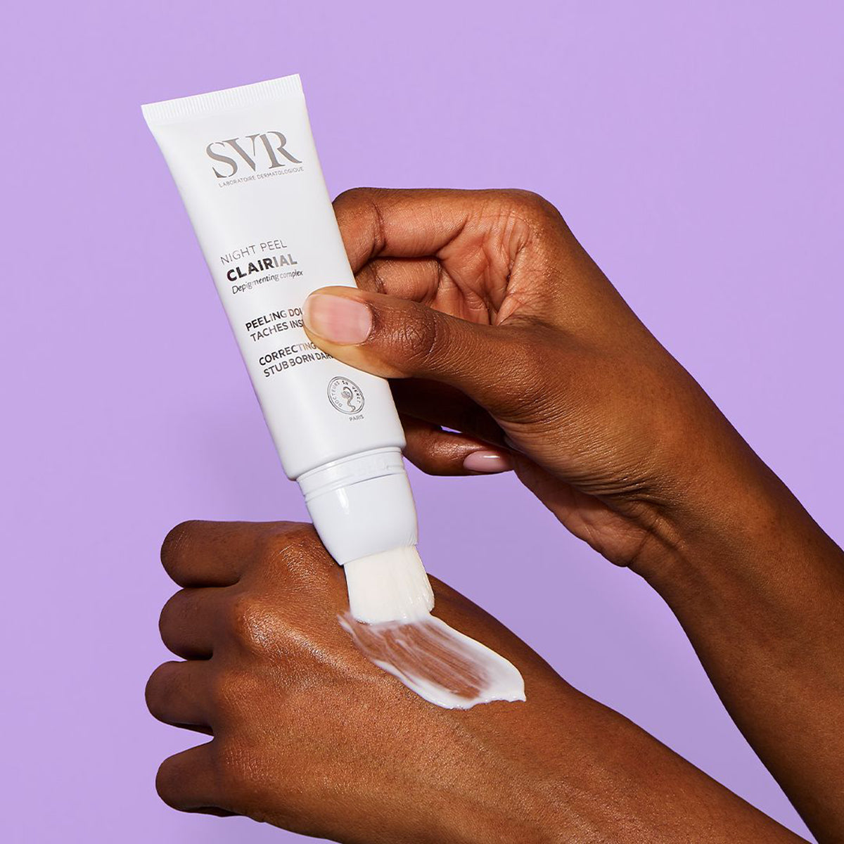 Tubo blanco de SVR Clairial Night Peel aplicando crema con su pincel integrado sobre la mano contra un fondo morado.