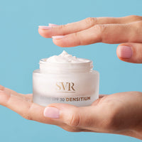 Manos de una mujer soseniendo en una de sus manos el envase del producto Creme SPF 30 Densitium de SVR en un fondo azul