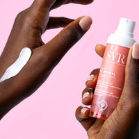 Manos de una mujer de tez oscura en una de sus manos sosteniendo el producto del Baume PALPEBRAL y en la otra mano aplicacion de la crema con fondo rosa