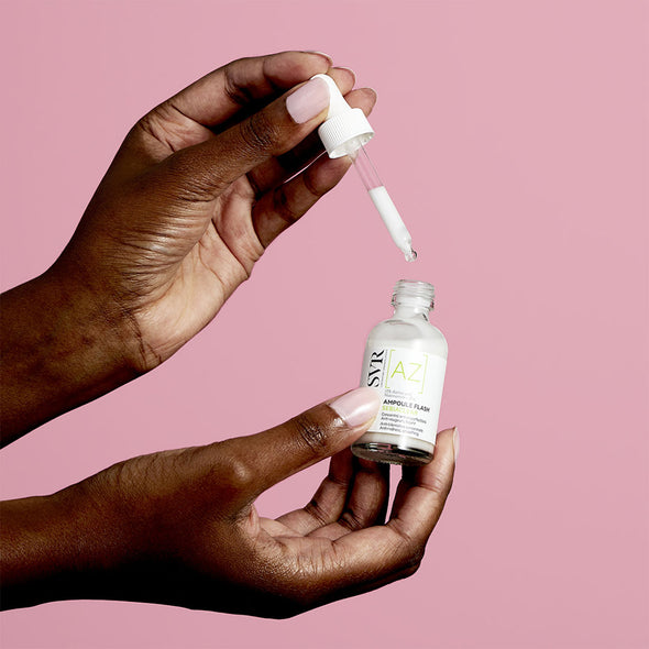 Manos de mujer de tez morena sosteniendo en una de sus manos un gotero y en otra mano la Ampoule Flash AZ de Sebiaclear de SVR con un fondo rosa