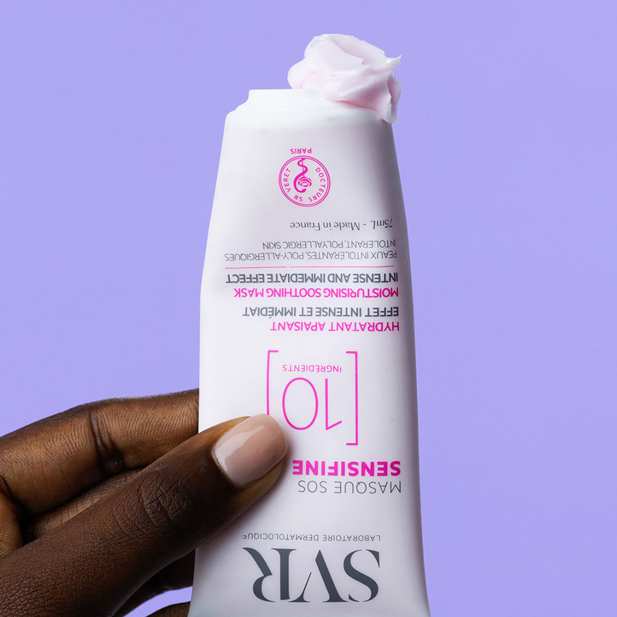 Mano de mujer de tez oscura sosteniendo el producto Sensifine Masque SOS en una de sus manos en un fondo morado claro