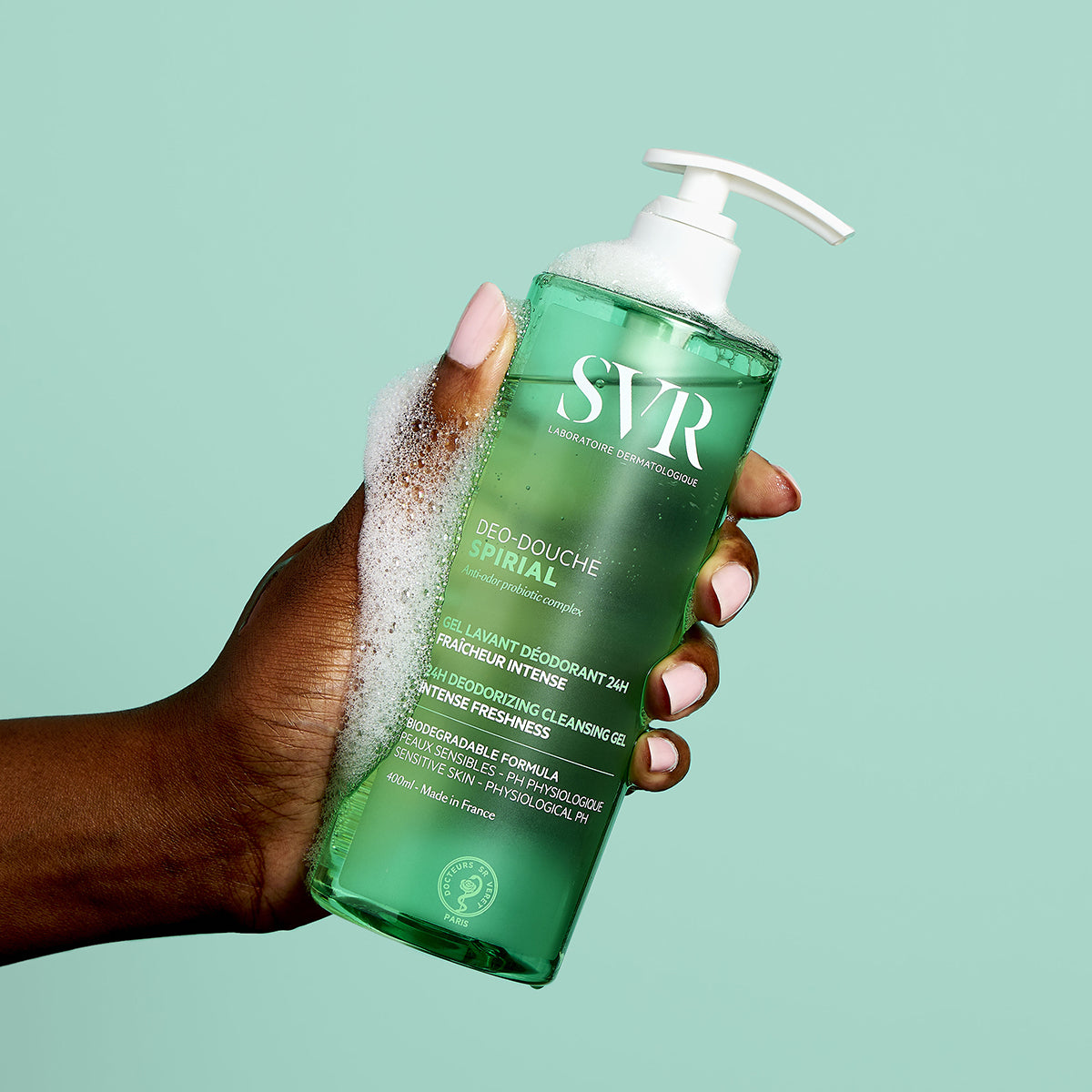 Una mano de tez oscura con uñas rosas, mostrando una botella del producto con jabon y espuma en la mano del Deo-Douche SPIRIAL gel limpiador y desodorante