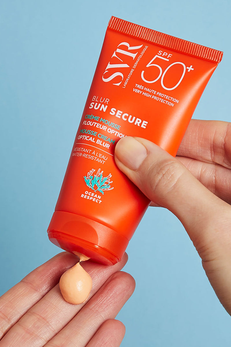 Manos de una mujer de tez clara aplicando en una de sus manos el Sunsecure Blur 50+SPF de SVR en un fondo azul