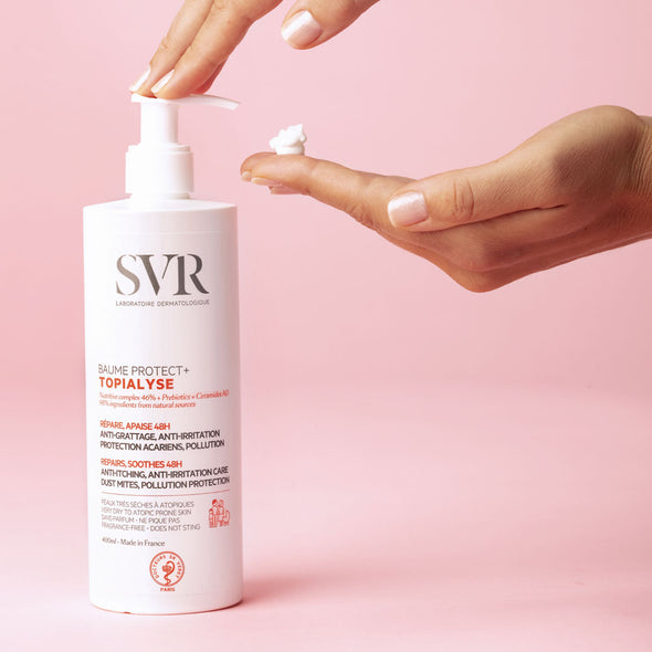Una mujer aplicandose en la mano con el dispensador el Baume protect+ TOPIALYSE, en fondo rosa, para pieles secas y sensibles.