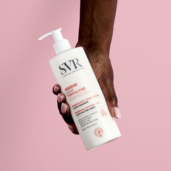 Producto envase de 400 ml dispensador de TOPIALYSE Crème en una mano de piel obscura con fondo rosa