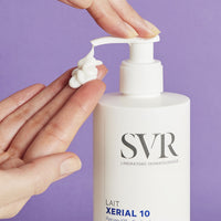 Un par de manos aplicando producto en una de sus manos de forma cremosa del dispensador del Xerial 10 Lait de SVR, con fondomorado