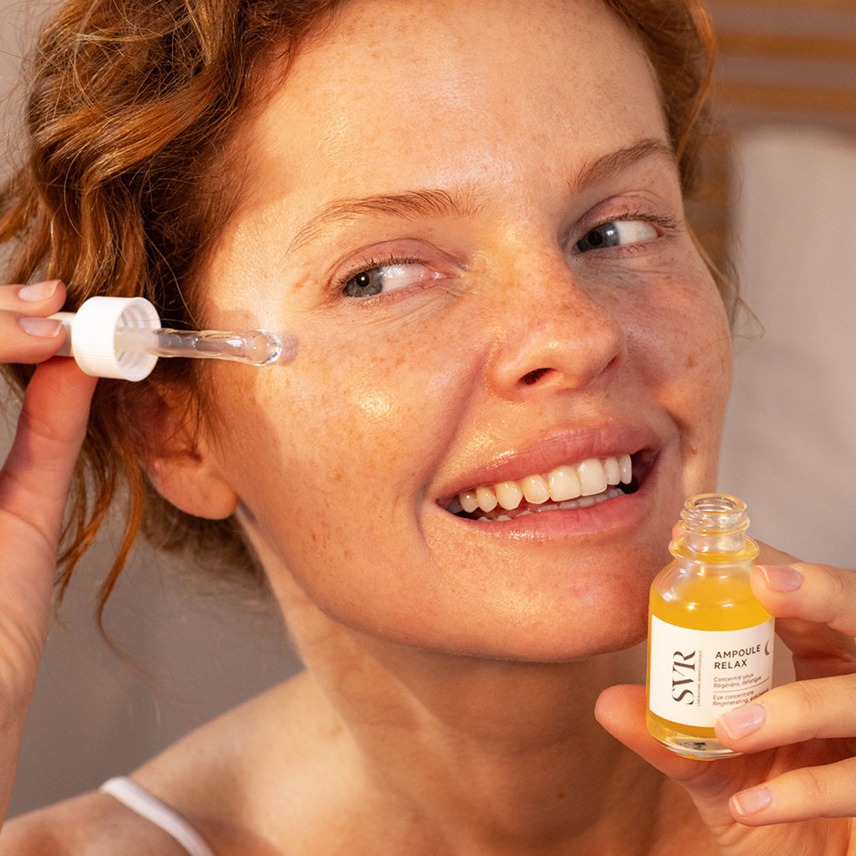 Mujer sonriente aplicando sérum nocturno SVR Ampoule Relax con gotero sobre el contorno de ojos en un frasco de cristal.