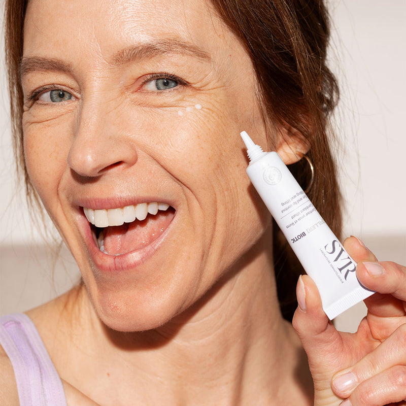 Una mujer de tez clara y piel madura con lineas de expresión sonriente, sosteniendo en una de sus manos con aplicacion de producto en una de sus mejillas del Filler Biotic de SVR en un fondo blanco