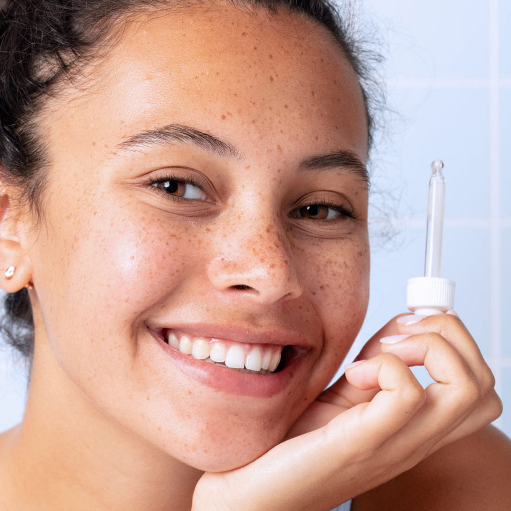 Una mujer de tez clara con pecas en el rostro sonriente, sosteniendo un gotero