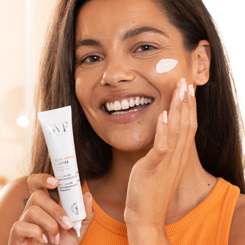 Mujer de tez morena claro sonriendo, con aplicación de producto en su mejilla y en la otra mano sosteniendo el producto Creme SPF50+ Clairial de SVR