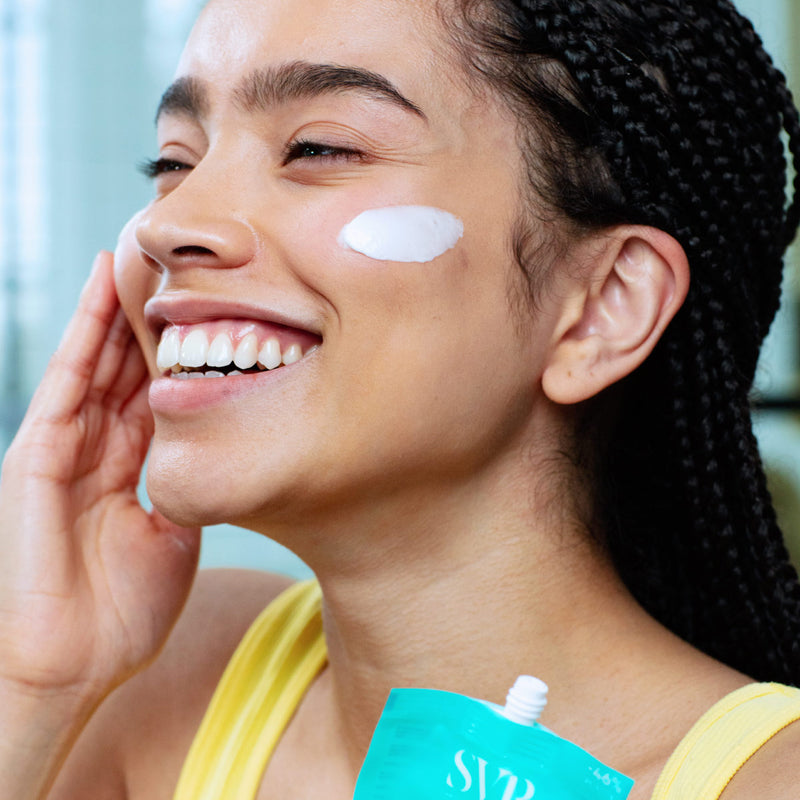 Mujer de tez morena de perfil sonriendo y con aplicación en una de sus mejilla del la Creme Hydraliane de SVR con un fondo claro