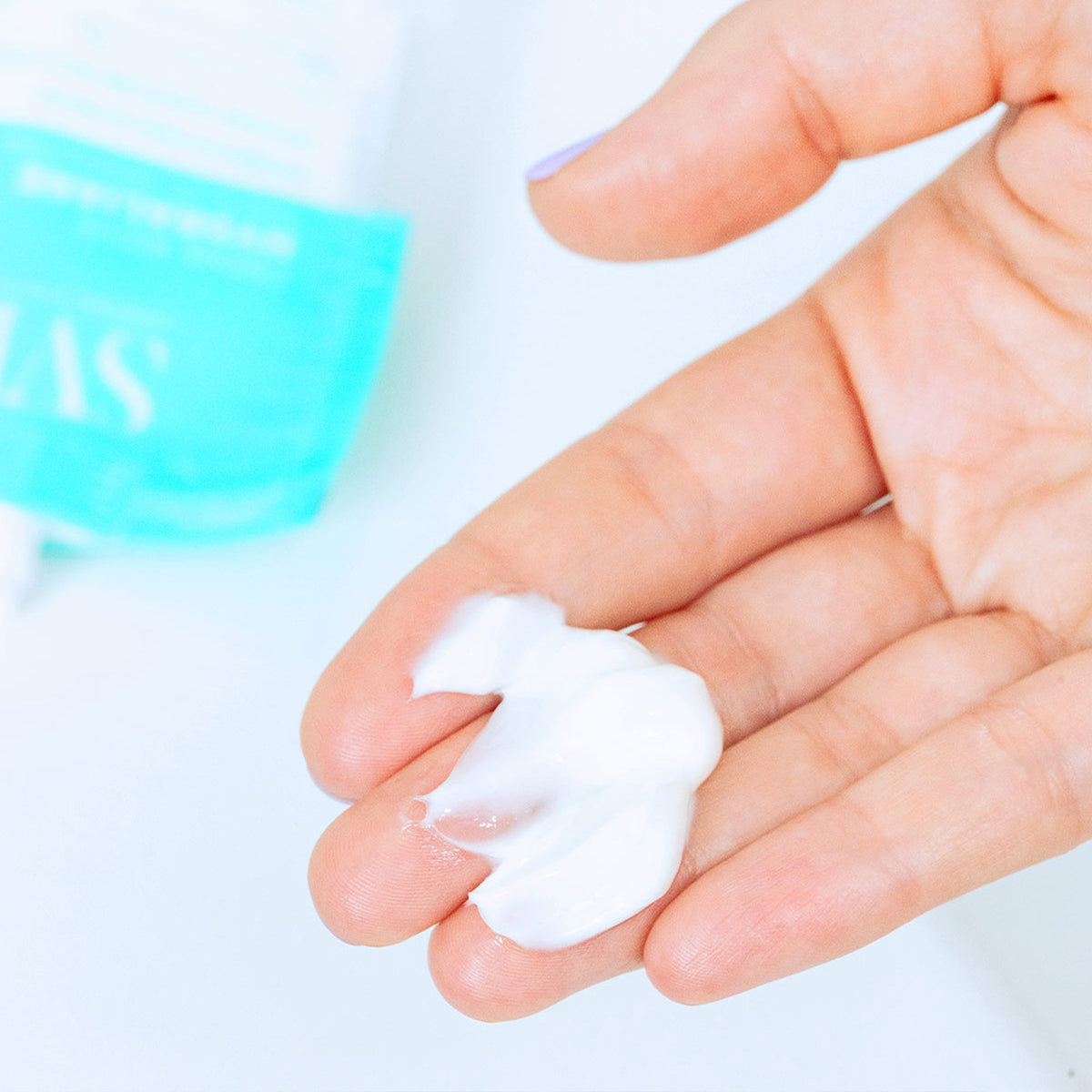 Textura cremosa blanca de Hydraliane Crème Riche sobre la palma de una mano con envase azul desenfocado al fondo.