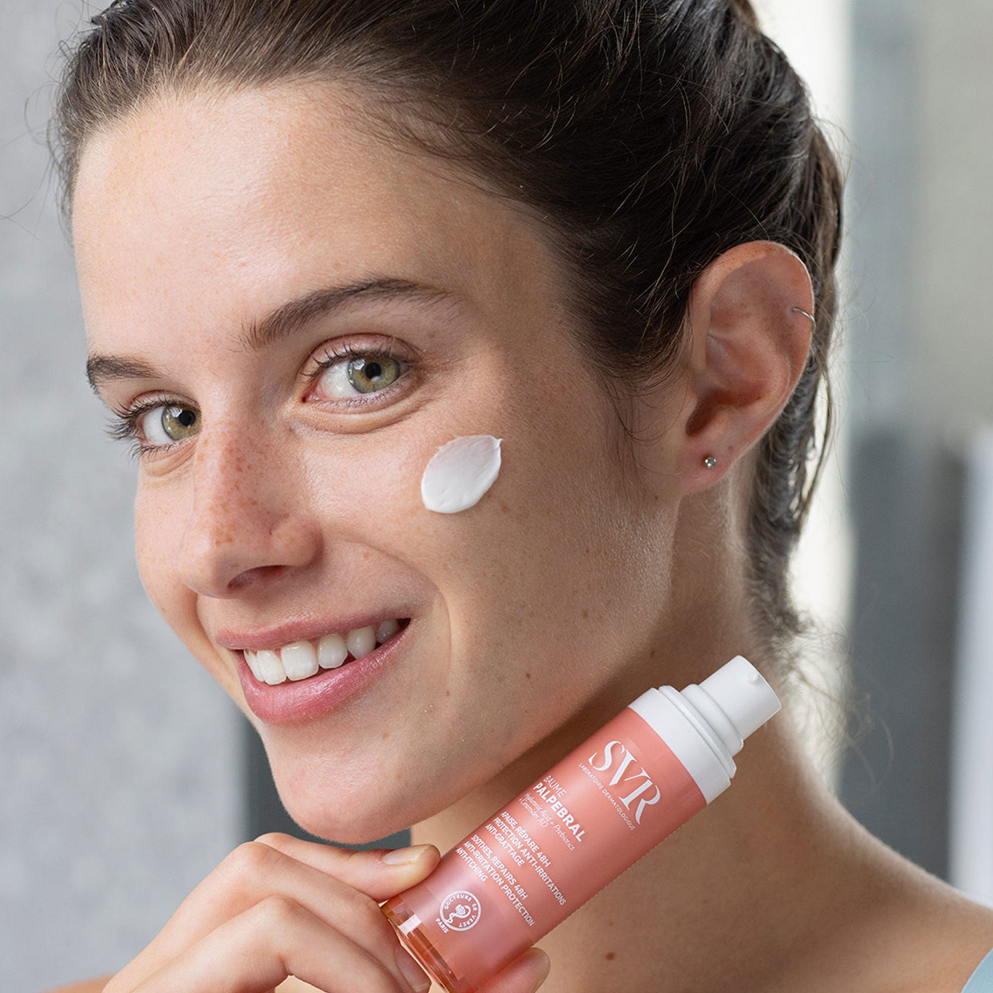 Mujer de tez clara cabello castaño ojos claros, sosteniendo el producto Baume PALPEBRAL con un poco de aplicacion en el rostro de crema blanca con fondo gris
