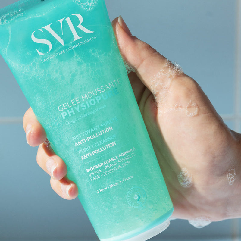 Una mano de tez clara mostrando el producto del gel PHYSIOPURE Gelee Moussante con espuma en la mano en un fondo azul