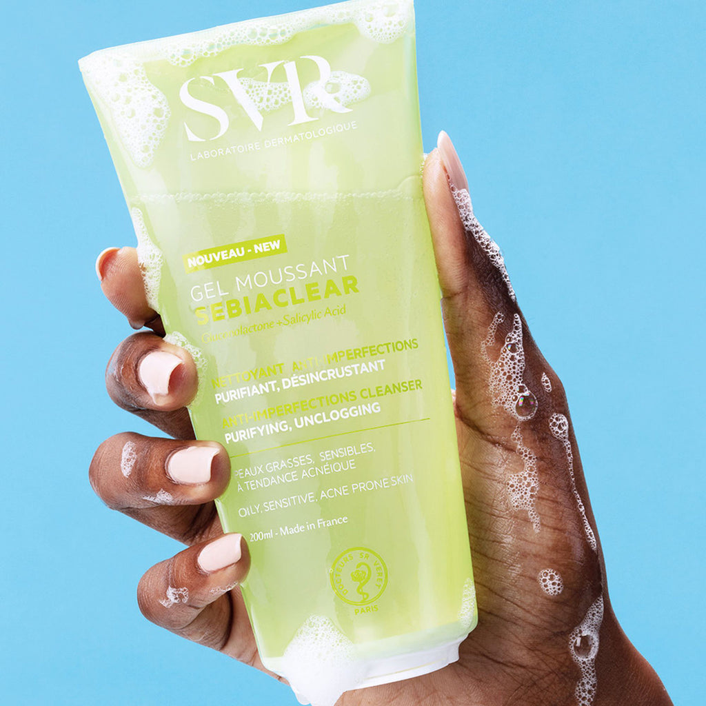 Una mano de tez obscura tomando la botella  de 200 ml del SEBIACCLEAR Gel Moussant de SVR con ligera espuma sobre la mano para pieles con tendencia acneica, en un fondo azul