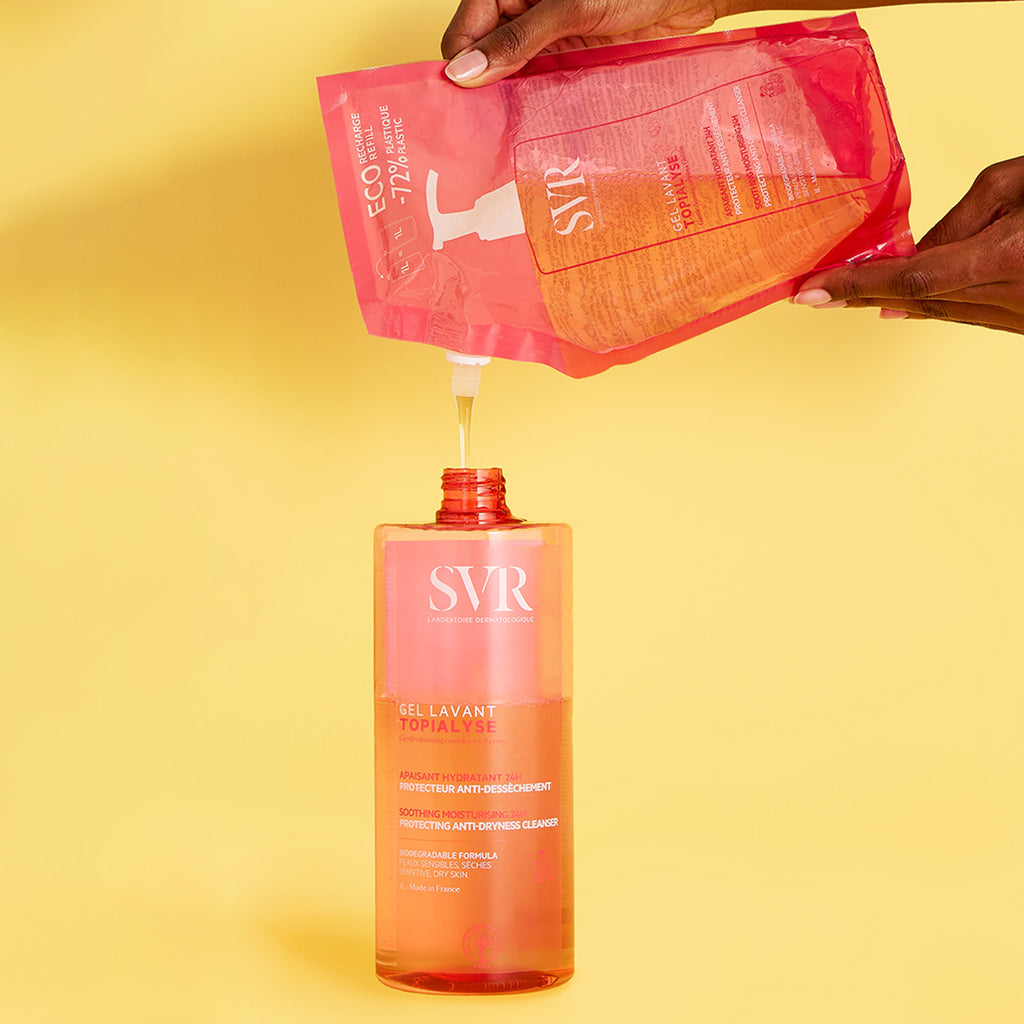 Unas manos de mujer rellenado una botella de Topialyse Gel Lavat de un empaque de refil en un fondo rosa