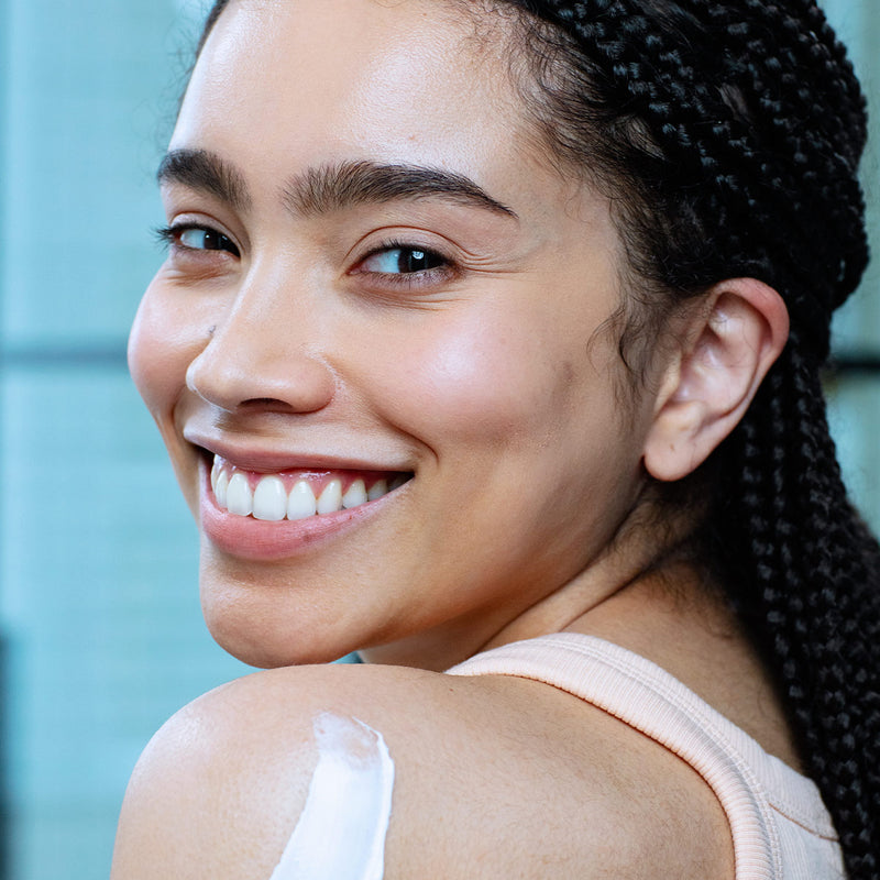 Rostro de mujer de tez morena con ojos grandes sonriendo, con producto aplicado en la parte del hombro como cremosa de color blanca, con fondoazul claro.