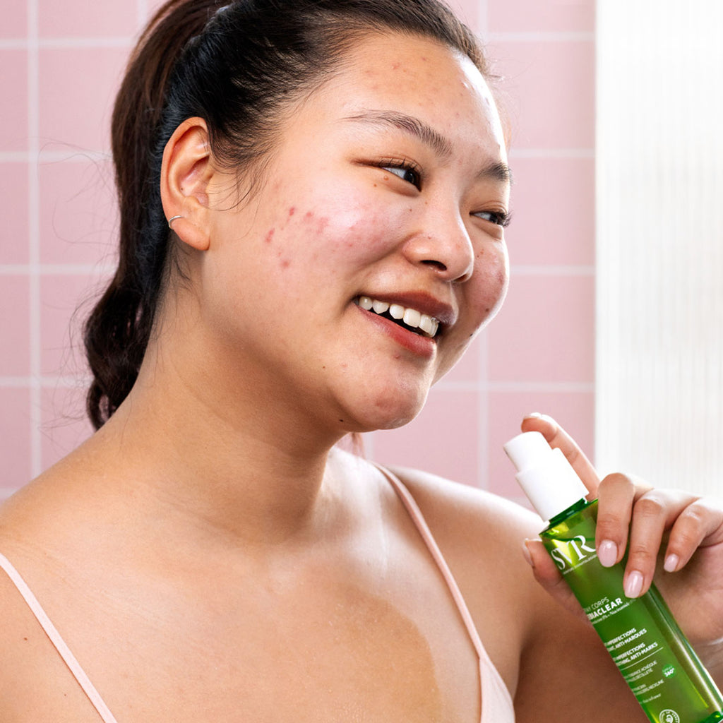 Mujer sonriendo de tez clara oriental con imperfecciones en el rostro sosteniendo  en una de sus manos el producto en su escote el spray corps de Sebiaclear de SVR