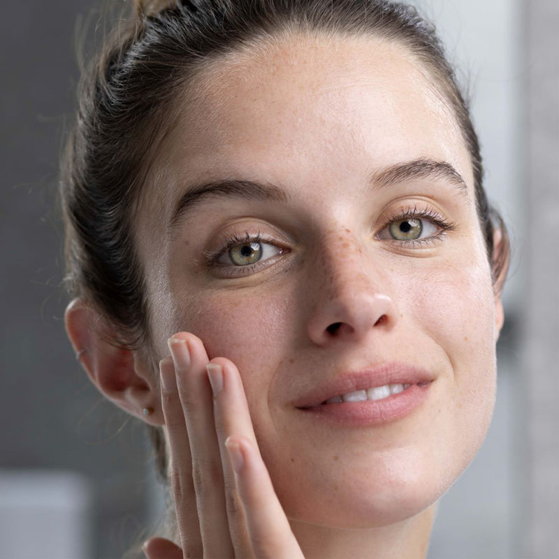 Una mujer de tez clara con cabello castaño con pecas en la nariz, tocando su mejilla como si se estuviera aplicando crema en la cara en un fondo gris