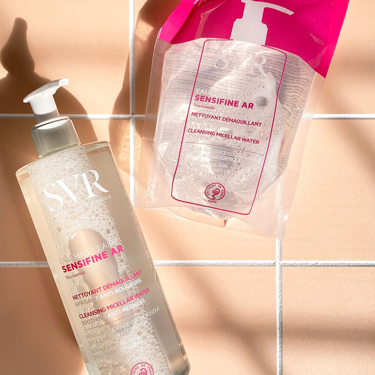 Muestra de productos EAU Micellaire SENSIFINE AR en presentacion de botella de 400 ml y eco refill para pieles sensibles con un fondo rosa claro y dandole el sol a los productos generando sombra de los productos.