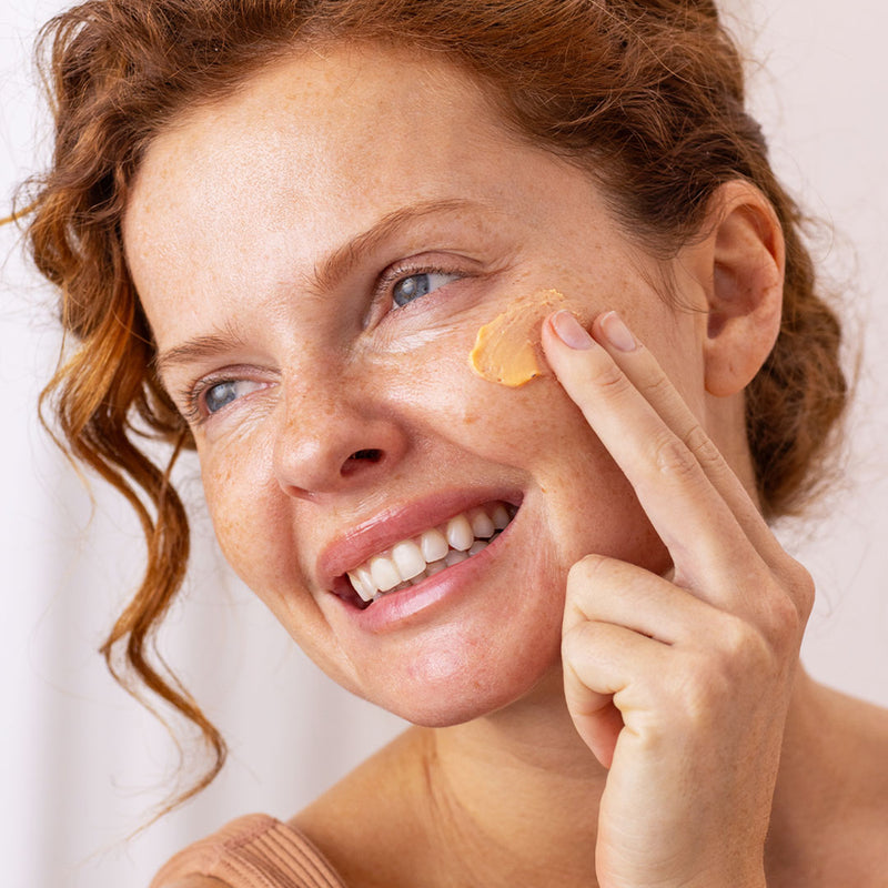 Una mujer de tez clara sonriendo de cabello pelirrojo con pecas en el rostro aplicando protector solar con tinte en el rostro en un fondo claro