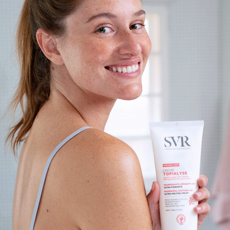 Mujer de cabello castaño claro con pecas y sonriendo en una mano esta agarrando una botella del SVR TOPIALYSE Crème perfecta para pieles secas y sensibles
