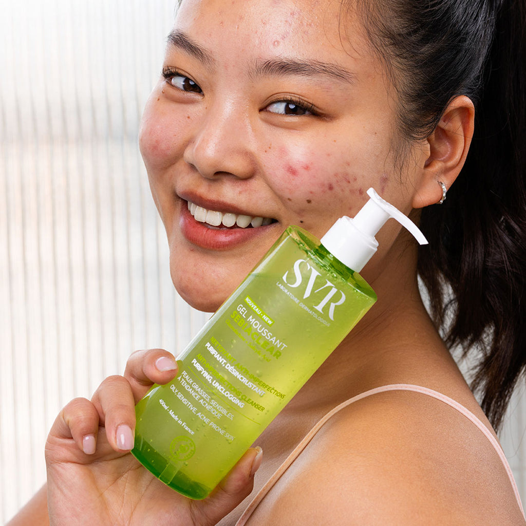 Mujer asiatica de cabello obscuro sonteniendo con una de sus manos mostrando a la altura de su rostro el producto SEBIACLEAR Gel Moussant de 400 ml con fondo blanco