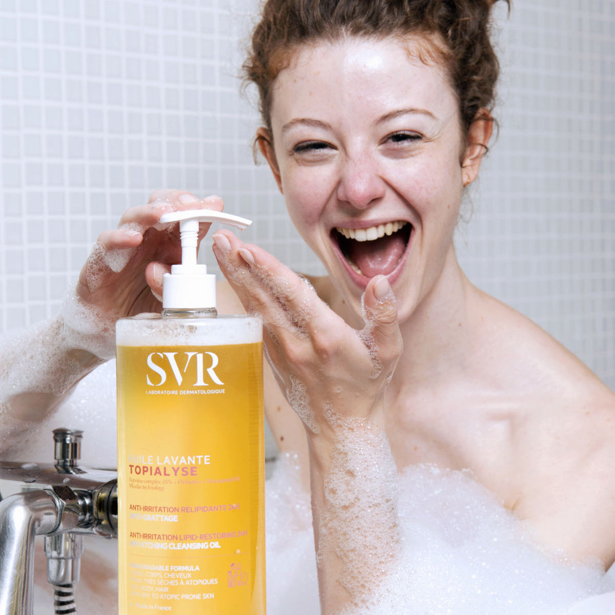 Mujer de tez clara con cabello rosado y sonriente aplicando producto del Topialyse Huile Lavante en una bañera con espuma y fondo blanco