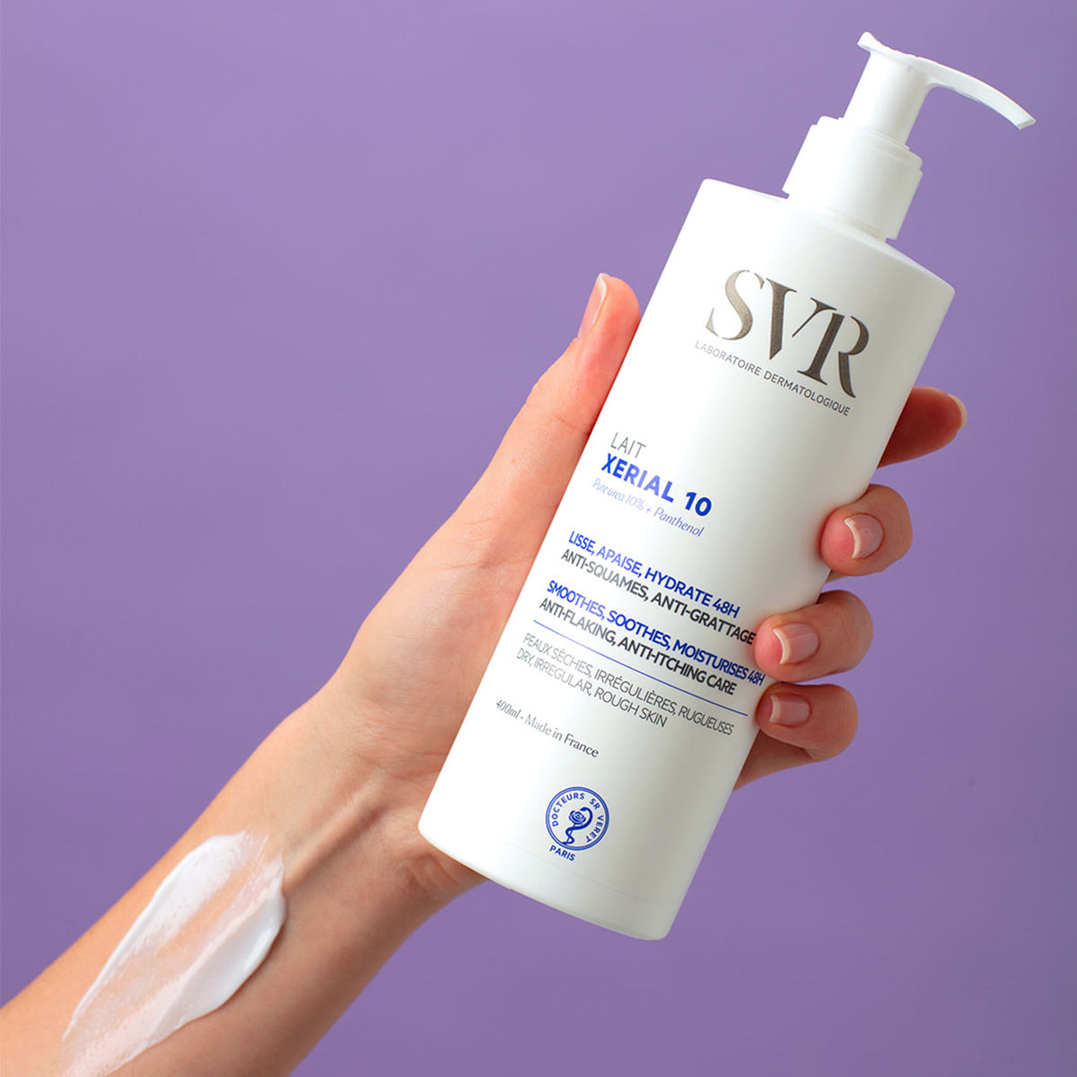 Un brazo de mujer tomando la botella del Lait XERIAL 10 de SVR, con un poco de producto enbarrado en la muñeca tipo crema blanca con fondo morado