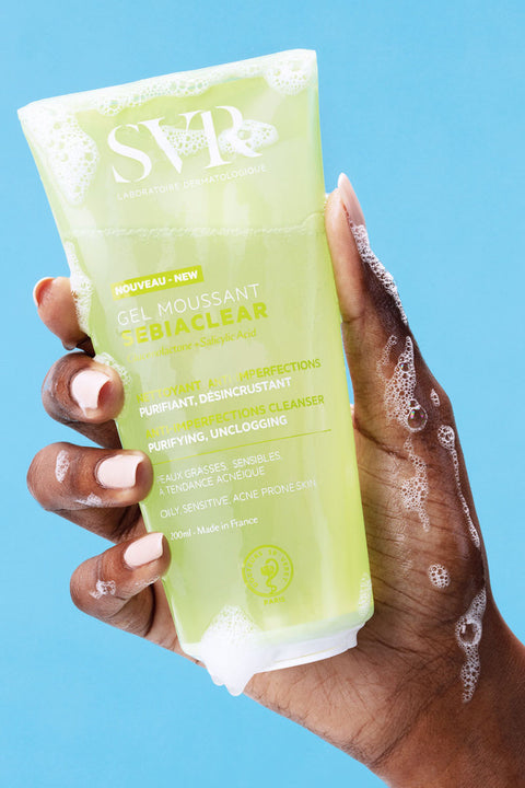 Una mano de tez obscura tomando la botella  de 200 ml del SEBIACCLEAR Gel Moussant de SVR con ligera espuma sobre la mano para pieles con tendencia acneica, en un fondo azul