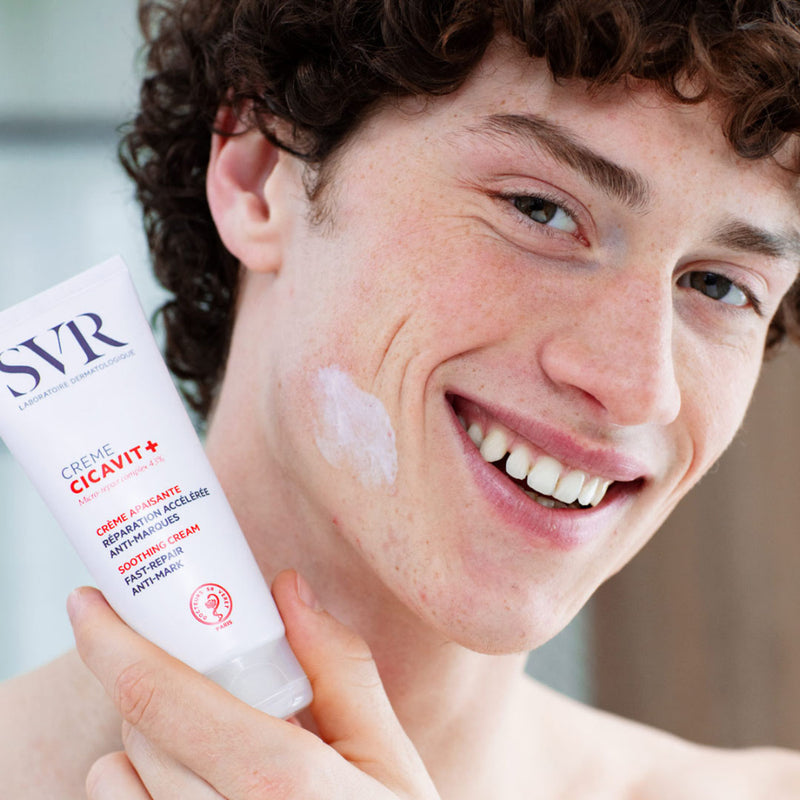 Hombre joven sonriendo mostrando en una de sus manos a la altura de su cuello el producto CICAVIT+ Creme con aplicacion en una de sus mejillas.