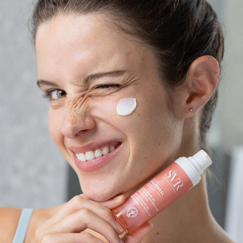 Mujer de tez clara cabello castaño ojos claros, sosteniendo el producto Baume PALPEBRAL con un poco de aplicacion en el rostro de crema blanca con fondo gris