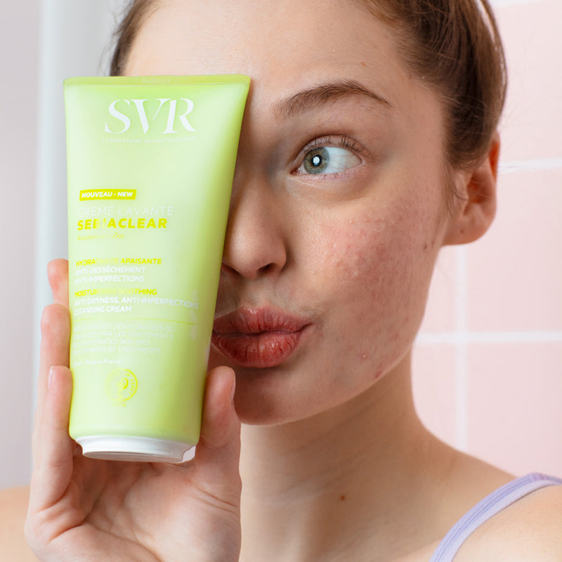 Una mujer de tez clara mostrando el producto con una de sus manos a la altura del rostro Creme Levante de SVR en presentación de 200 ml en un fondo tipo pared de baño color rosa