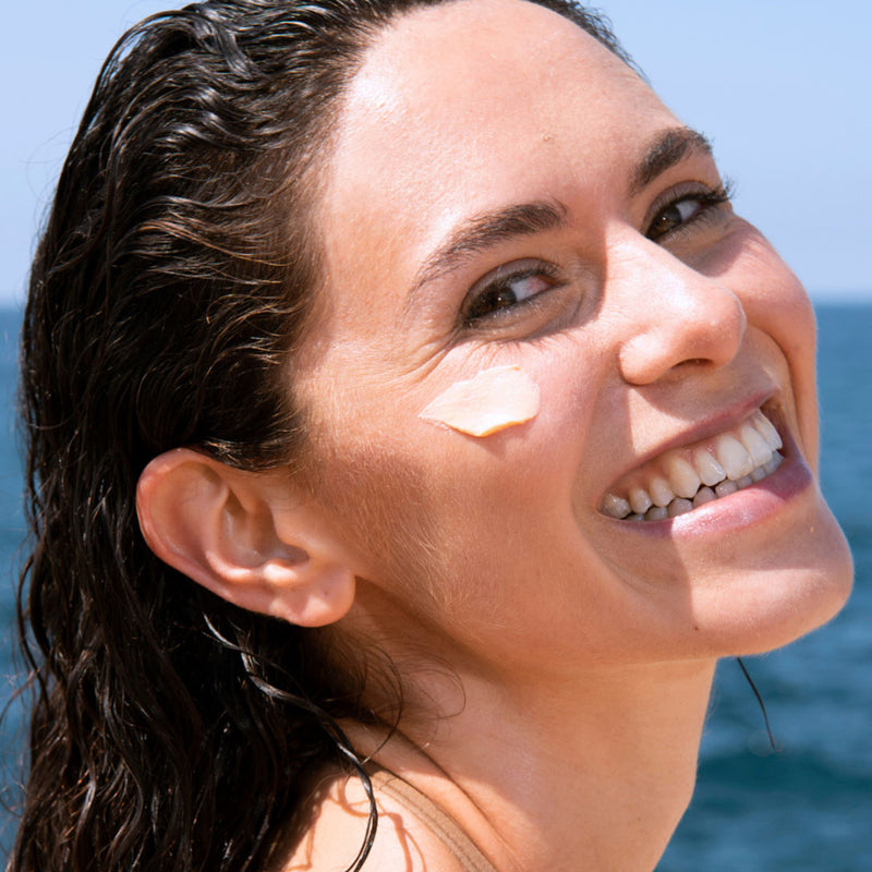 Una mujer de tez clara sonriendo con el pelo mojado en el mar, con aplicacion de protector solar en una de sus mejillas.