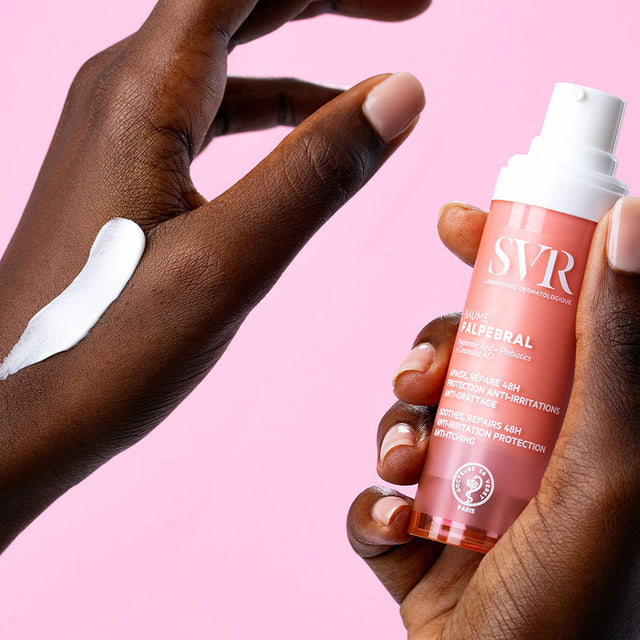 Manos de una mujer de tez oscura en una de sus manos sosteniendo el producto del Baume PALPEBRAL y en la otra mano aplicacion de la crema con fondo rosa