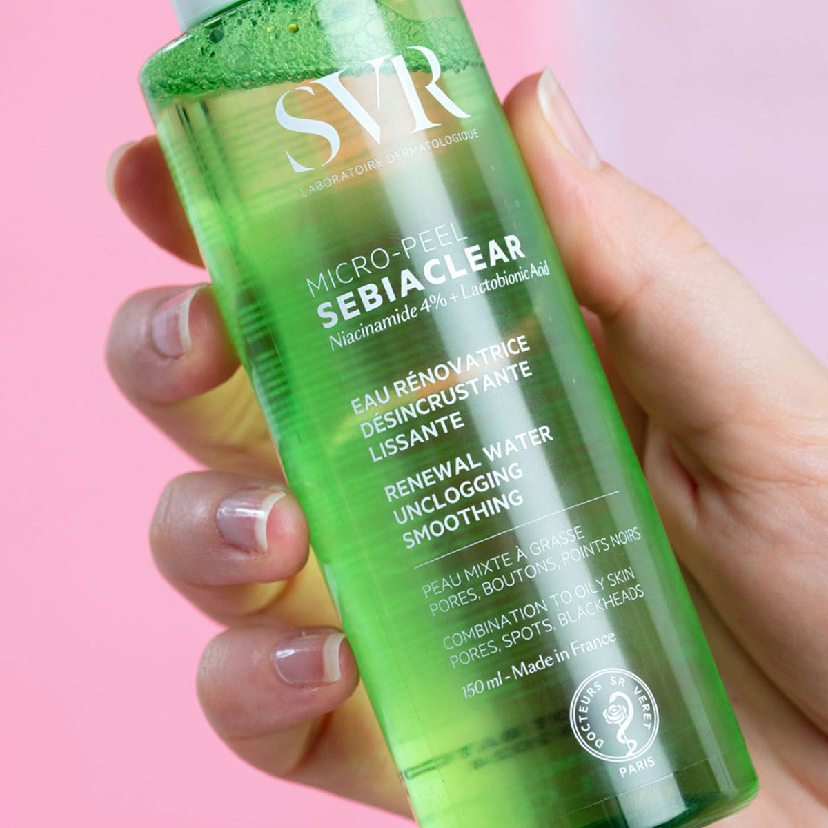 Botella transparente de 150ml de tónico SVR Sebiaclear Micro-Peel con líquido verde, sostenida en una mano sobre fondo rosa.