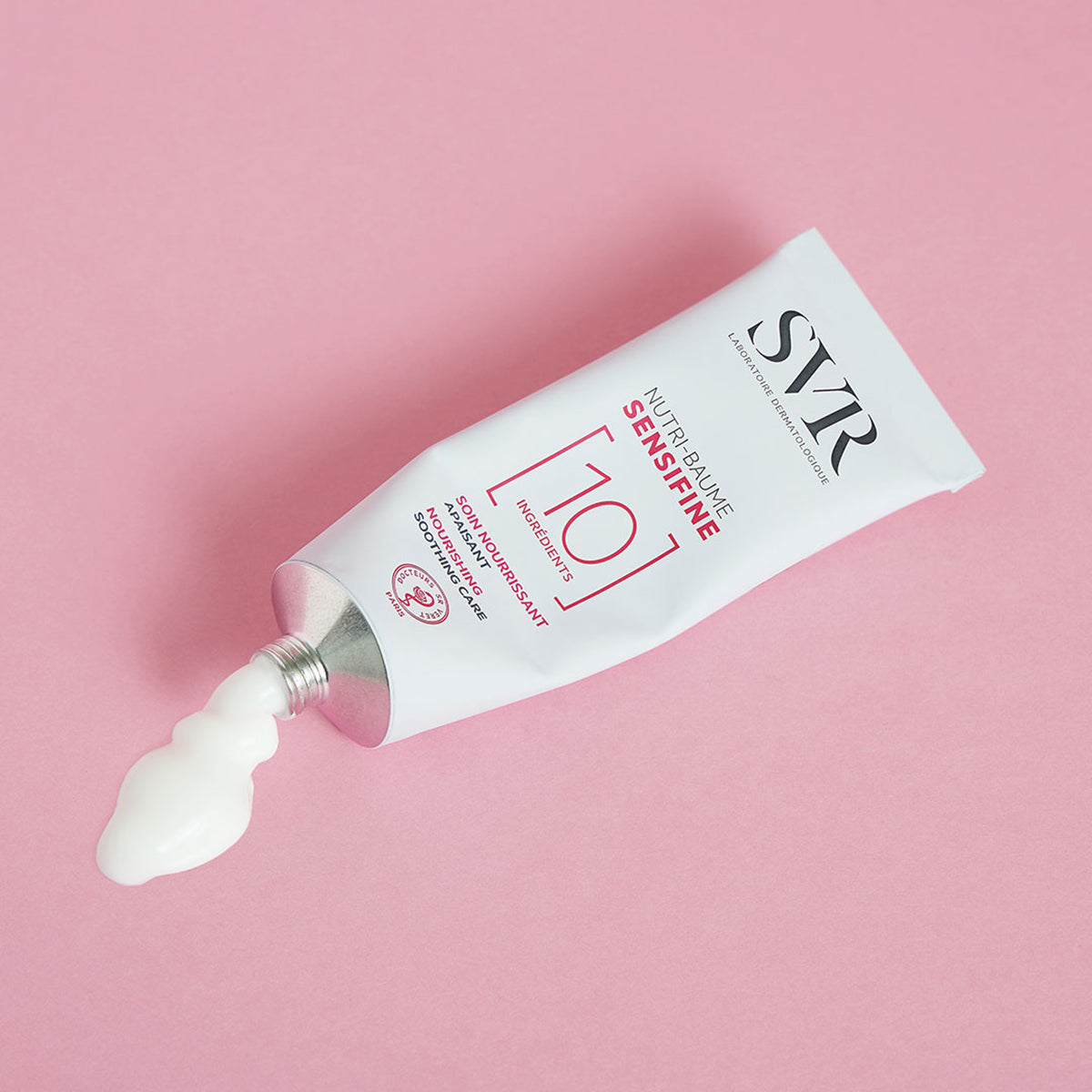 Tubo blanco de bálsamo SVR Sensifine Nutri-Baume con crema hidratante saliendo por la boquilla sobre fondo rosa.
