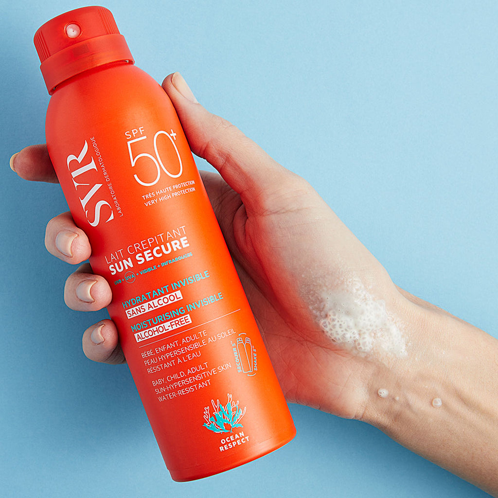 Mano de una mujer sosteniendo Sun Secure Lait Crepitant de botella naranja en fondo azul.