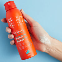 Mano de una mujer sosteniendo Sun Secure Lait Crepitant de botella naranja en fondo azul.