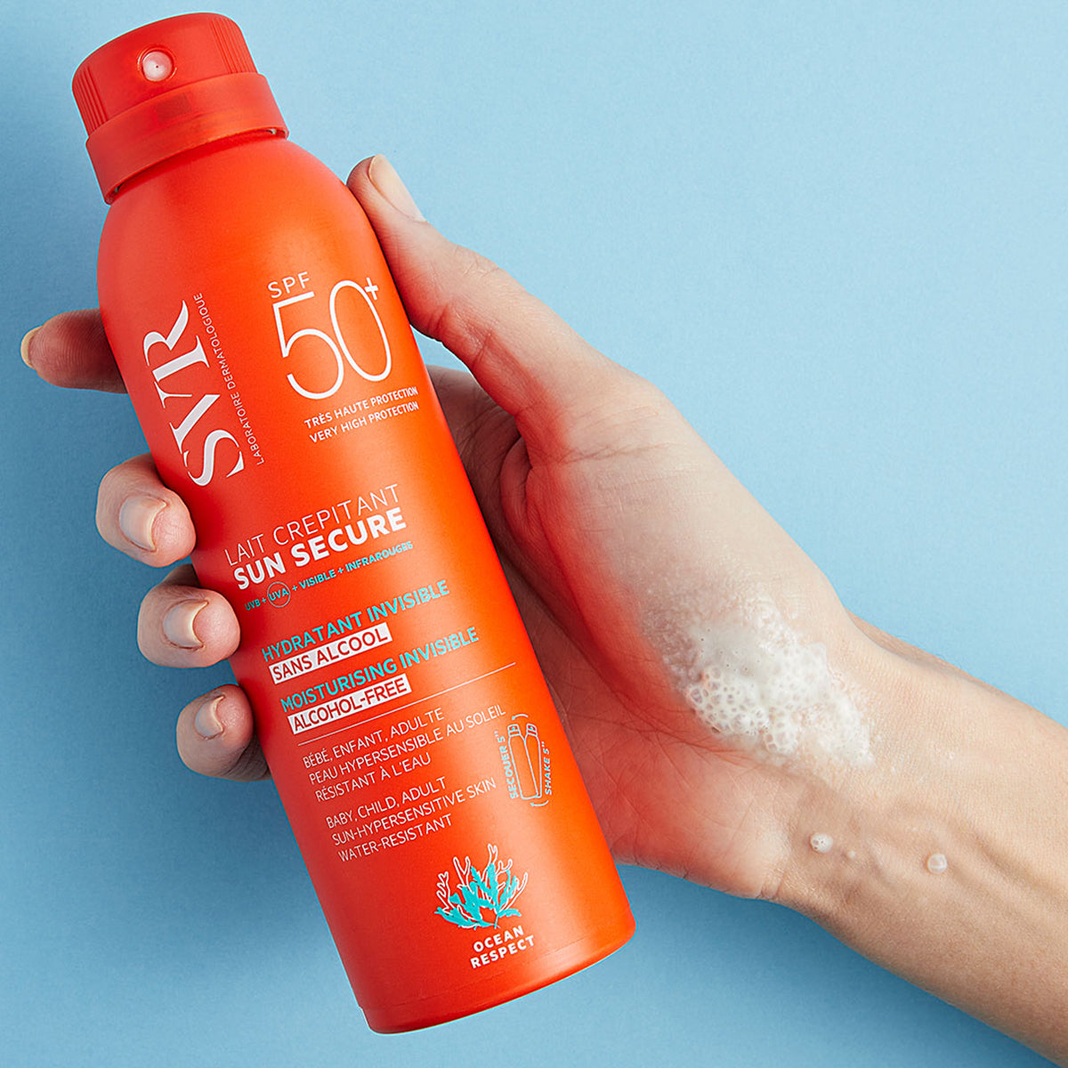 Mano de una mujer sosteniendo Sun Secure Lait Crepitant de botella naranja en fondo azul.