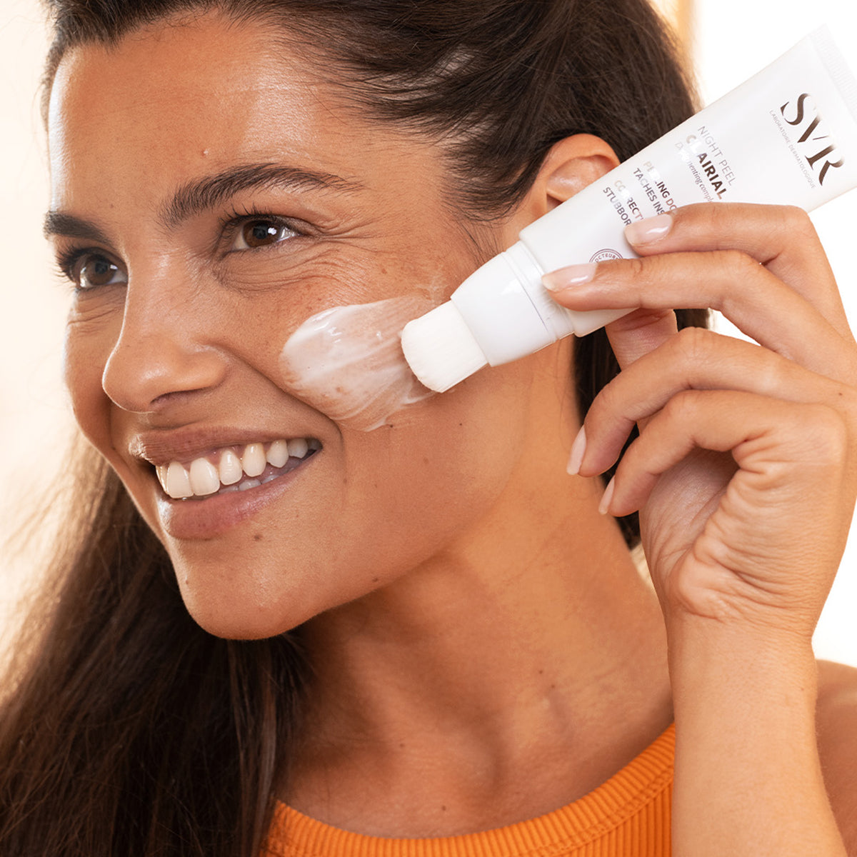 Mujer sonriente aplicando SVR Clairial Night Peel con el pincel integrado sobre su mejilla para reducir manchas.