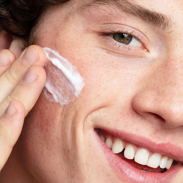 El rostro de un hombre joven sonriendo aplicando crema en una de sus mejillas