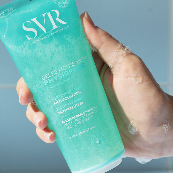 Una mano de tez clara mostrando el producto del gel PHYSIOPURE Gelee Moussante con espuma en la mano en un fondo azul