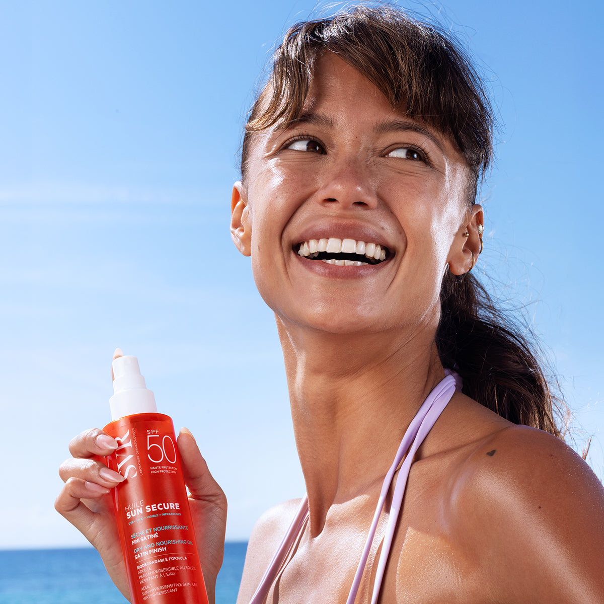 Mujer sonriente bajo el sol sosteniendo un frasco naranja de protector solar SVR SUN SECURE Huile SPF50 con acabado satinado.