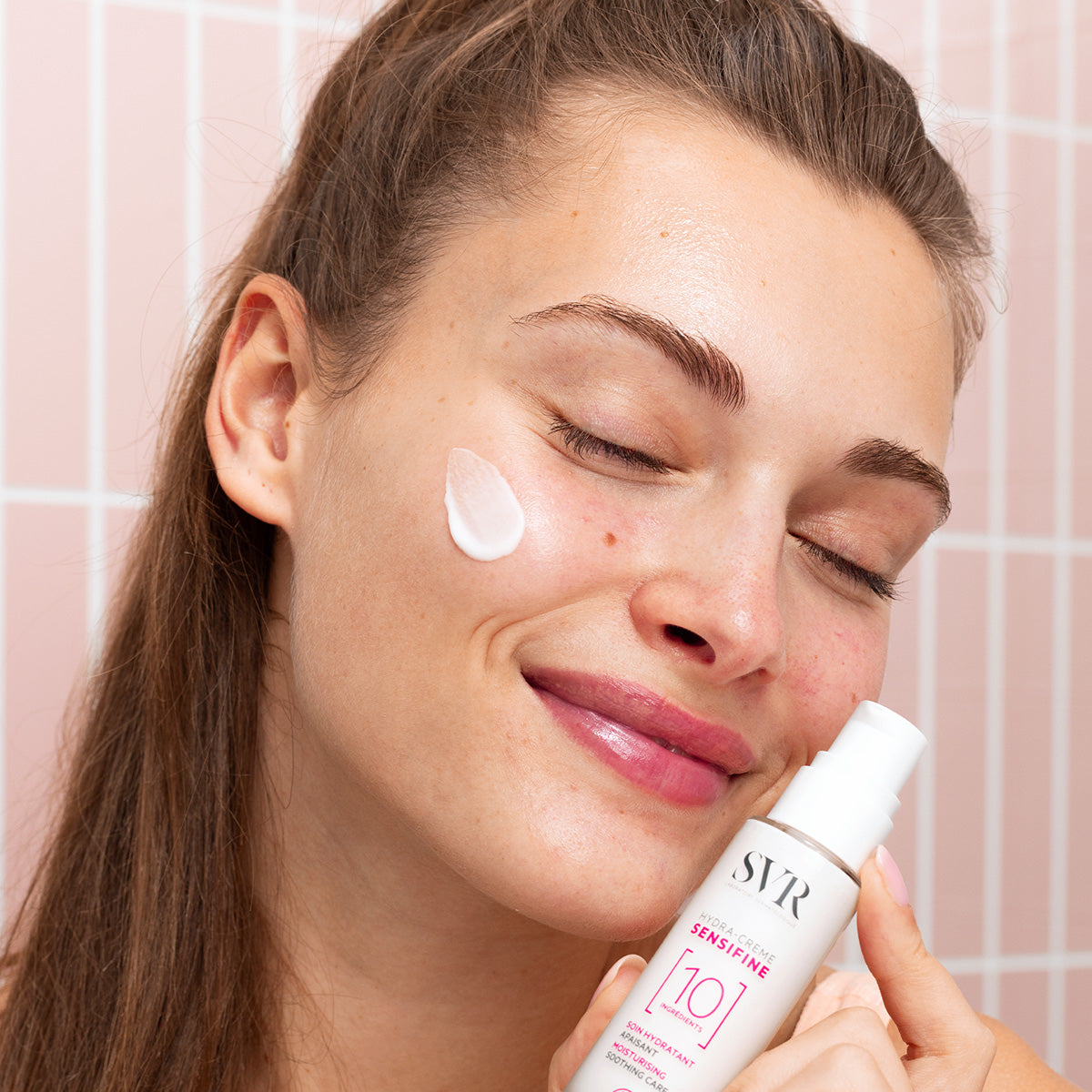 Mujer sonriente aplicando crema hidratante SVR Sensifine Hydra-Crème blanca en su mejilla frente a azulejos rosados.