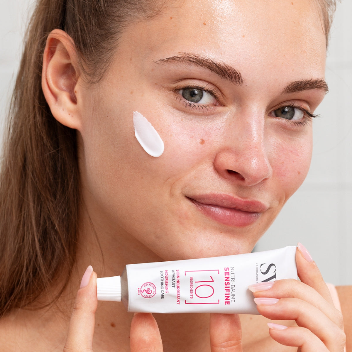 Mujer aplicando bálsamo hidratante SENSIFINE Nutri-Baume de SVR en su mejilla, sosteniendo el tubo blanco con texto rosa.