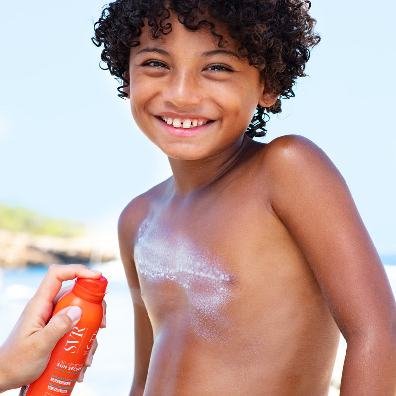 Mano de una mujer aplicando Sun Secure Lait Capritan en el pecho de un niño moreno con rizos. 