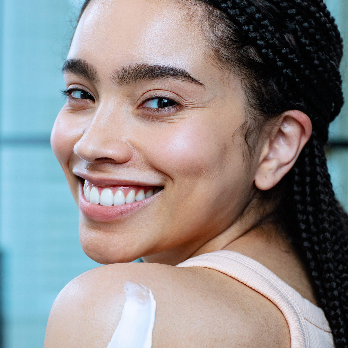 Rostro de mujer de tez morena con ojos grandes sonriendo, con producto aplicado en la parte del hombro como cremosa de color blanca, con fondoazul claro.