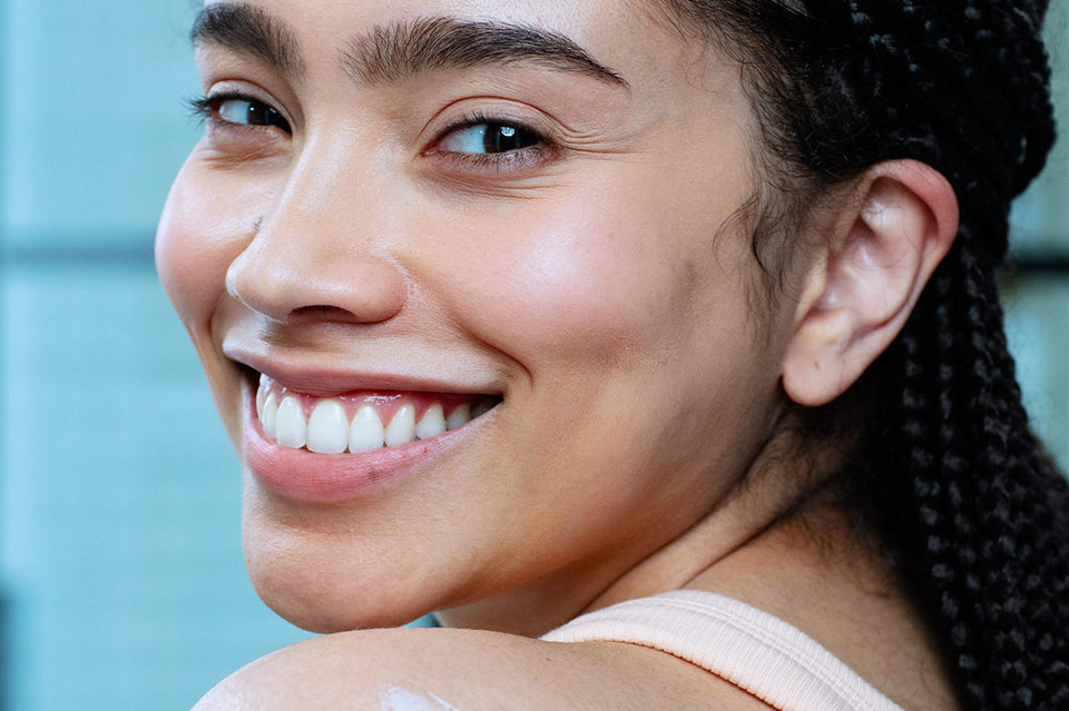 Rostro de mujer de tez morena con ojos grandes sonriendo, con producto aplicado en la parte del hombro como cremosa de color blanca, con fondoazul claro.